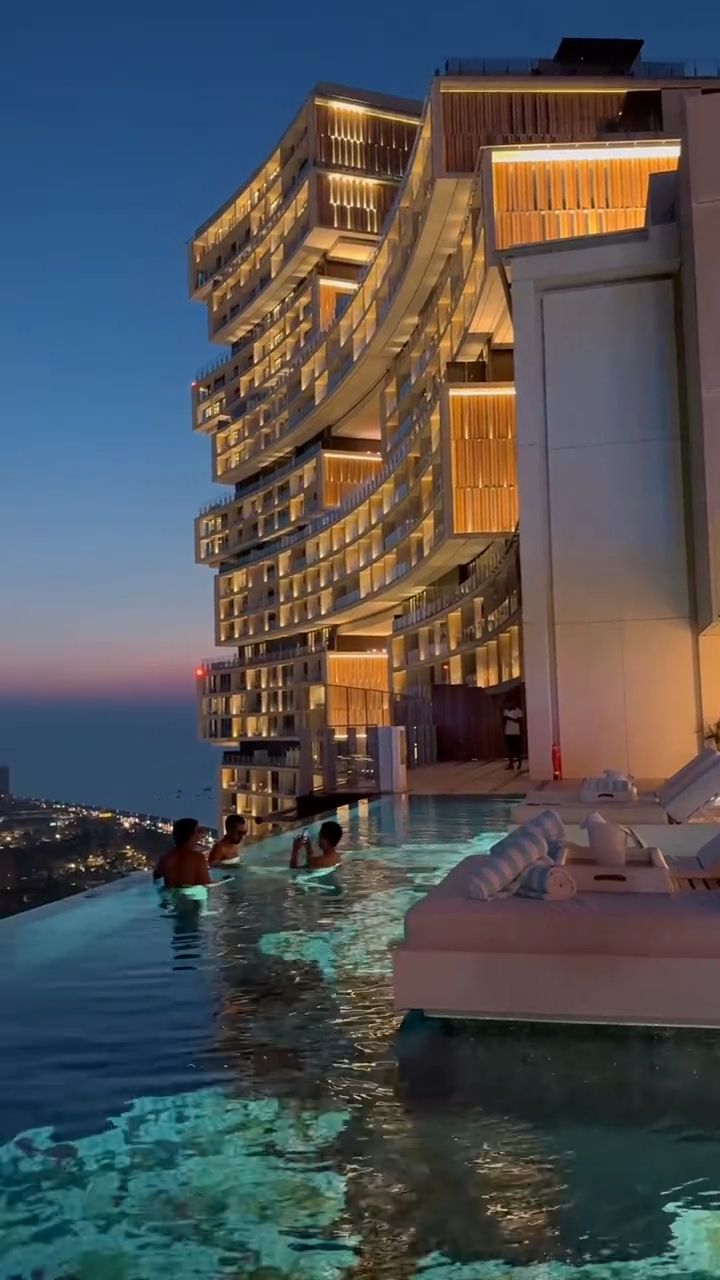 people are swimming in the pool next to an ocean side hotel at night time with lights on