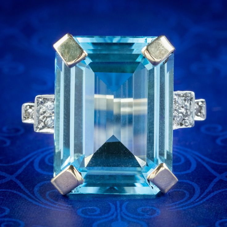 An outstanding Art Deco style cocktail ring boasting an impressive emerald cut blue topaz weighing approx. 26 carats. It has a clear ocean blue hue, similar to that of an aquamarine and is complemented by three twinkling brilliant cut diamonds on each shoulder.  The topaz is held securely in a pronounced 9ct gold claw mount which crowns it high upon the finger. It's an eye-catching statement piece and complete with Birmingham hallmarks within the band.  We recommend you insure for £1,700. We can Modern Blue Baguette Cut Emerald Ring, Formal Light Blue Topaz Ring, Elegant Blue Rectangular Emerald Ring, Formal Blue Emerald Ring With Accent Stones, Rectangular Blue Topaz Ring With Prong Setting, Art Deco Emerald Cut Topaz Ring With Accent Stones, Classic Blue Octagon Topaz Ring, Art Deco Diamond Cut Topaz Ring For Formal Occasions, Art Deco Formal Topaz Ring With Diamond Cut