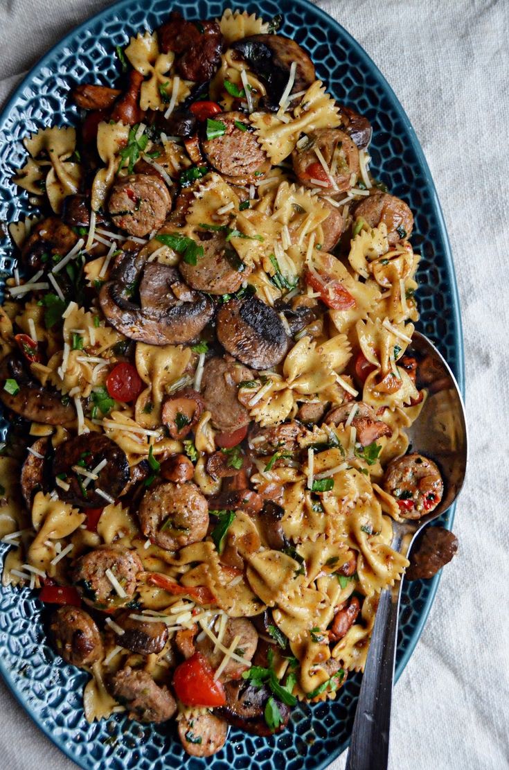 pasta with sausage, mushrooms and tomatoes in a blue bowl next to a spoon on a white table cloth