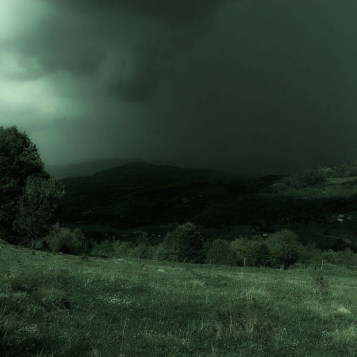 a green field with trees and mountains in the background under a dark sky filled with clouds