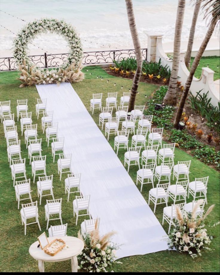 an outdoor ceremony setup with white chairs and flowers on the grass near the beach, surrounded by palm trees