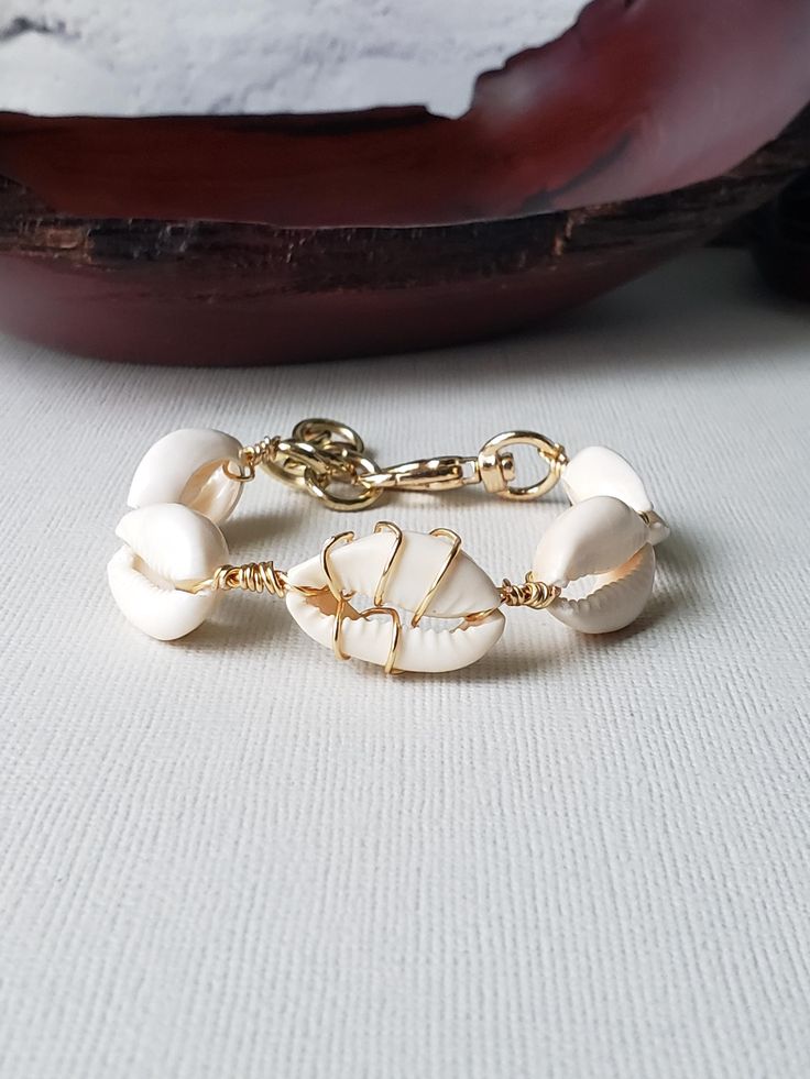 A sultry bracelet with the prettiest cowries. Center cowry is wire wrapped for interest. Done in your choice of gold or silver plated wire. Bold toggle clasp, XL ivory shells, and thick jumprings for extenders make this bracelet stand out. Fits from a 6.5" to 8.5" wrist.  Comes boxed and ready for gifting. Handmade White Shell-shaped Bracelets, White Cowrie Shell Bracelet, Elegant White Cowrie Shell Jewelry, White Hand Wrapped Bracelet, Unique Hand Wrapped White Bracelet, Unique Gold Jewelry For Vacation, White Bohemian Jewelry With Oyster Bracelet, Adjustable Cowrie Shell Bracelet Jewelry, Adjustable Cowrie Shell Bracelet