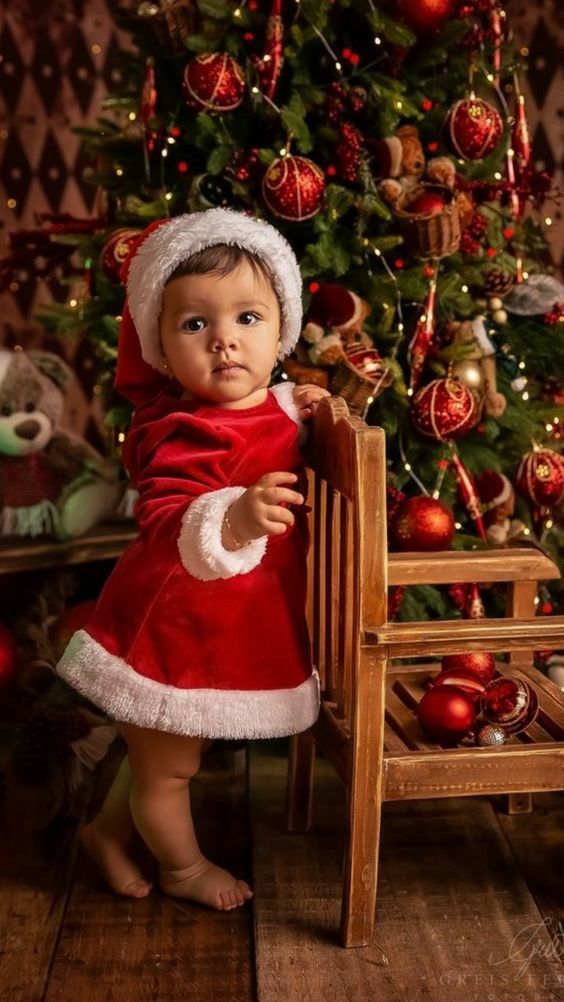 a baby dressed as santa claus standing in front of a christmas tree