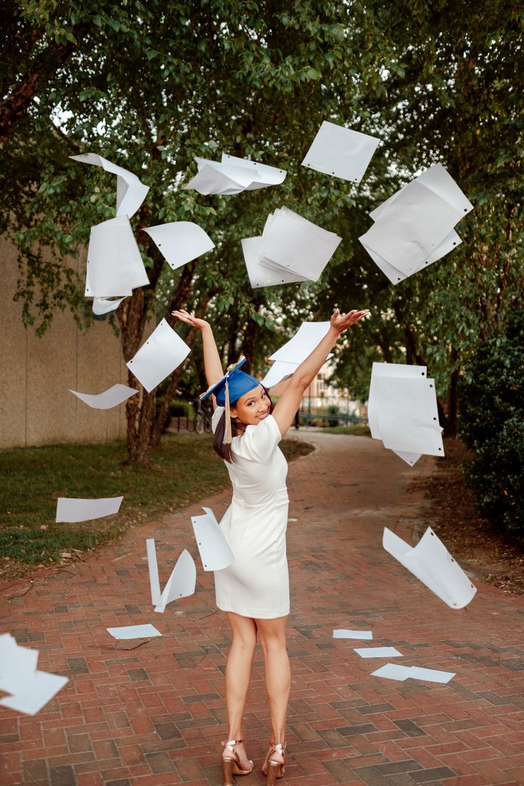 a woman in a white dress throwing papers into the air on a brick walkway with trees and bushes behind her