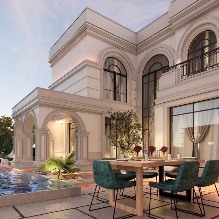 an outdoor dining area in front of a large white house with pool and fountain at dusk