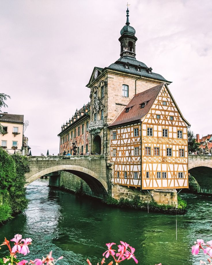 an old building next to a river with flowers in the foreground and a bridge crossing over it