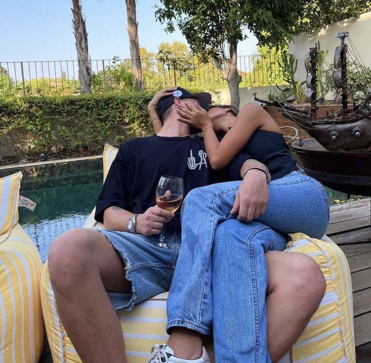 a man and woman sitting on top of each other next to a pool with a boat in the background