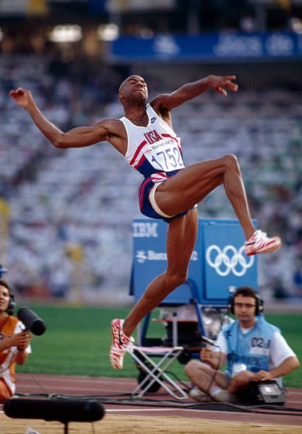 a man jumping in the air on top of a track with people watching behind him