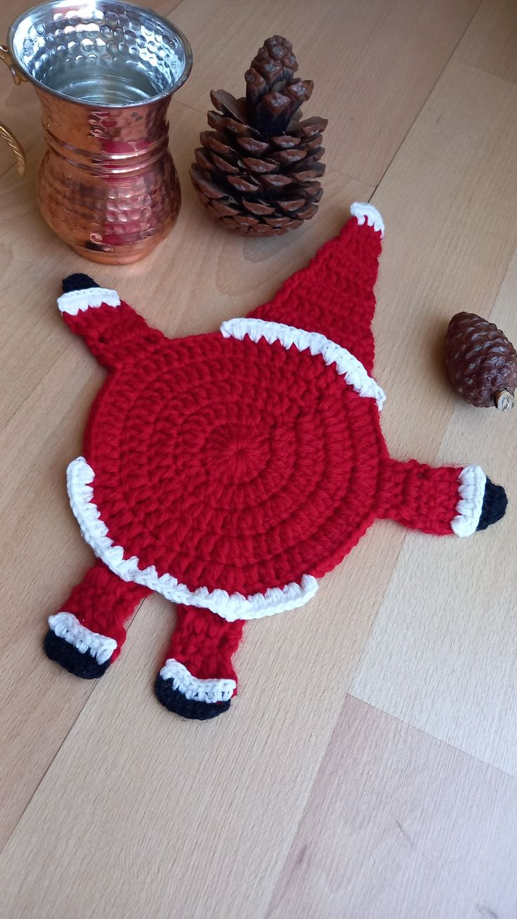 a crocheted red and white santa claus hat next to pine cones on a wooden table