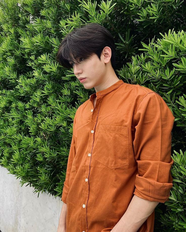 a young man standing in front of a bush wearing an orange shirt and black pants