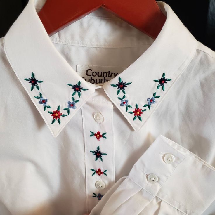 a white shirt with red, green and blue flowers embroidered on the collar is being held by a wooden hanger