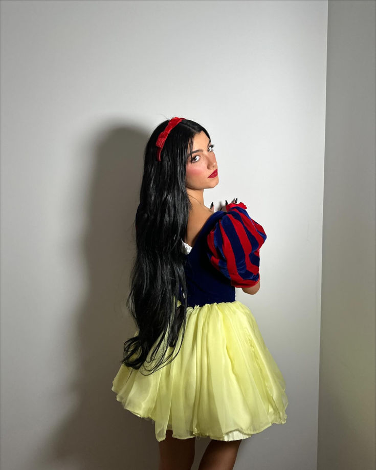 a woman with long black hair wearing a yellow skirt and red striped shirt is standing in front of a white wall