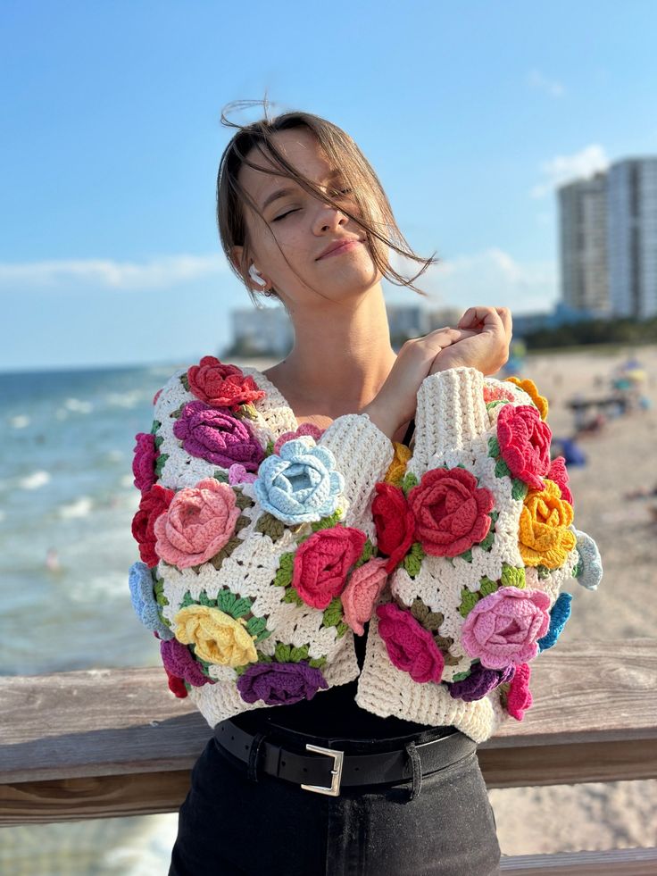 a woman standing on the beach wearing a crocheted sweater with roses in it