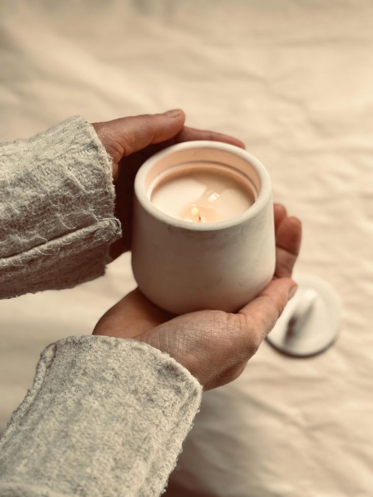 a person holding a white cup with a candle in it's hand on a bed