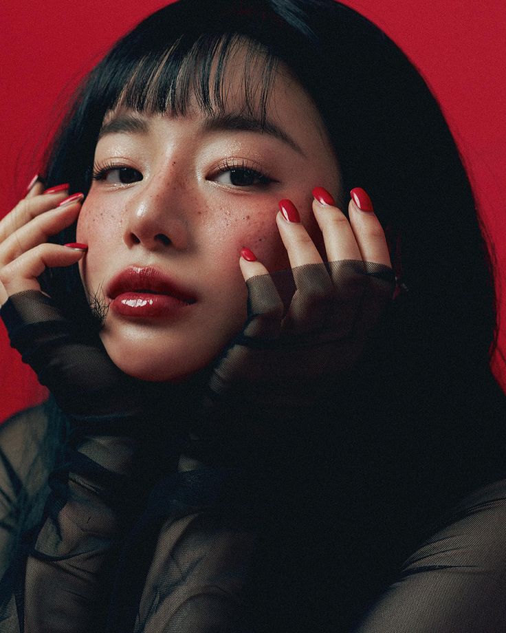 a woman with dark hair and red nail polish holding her hands to her face, against a red background