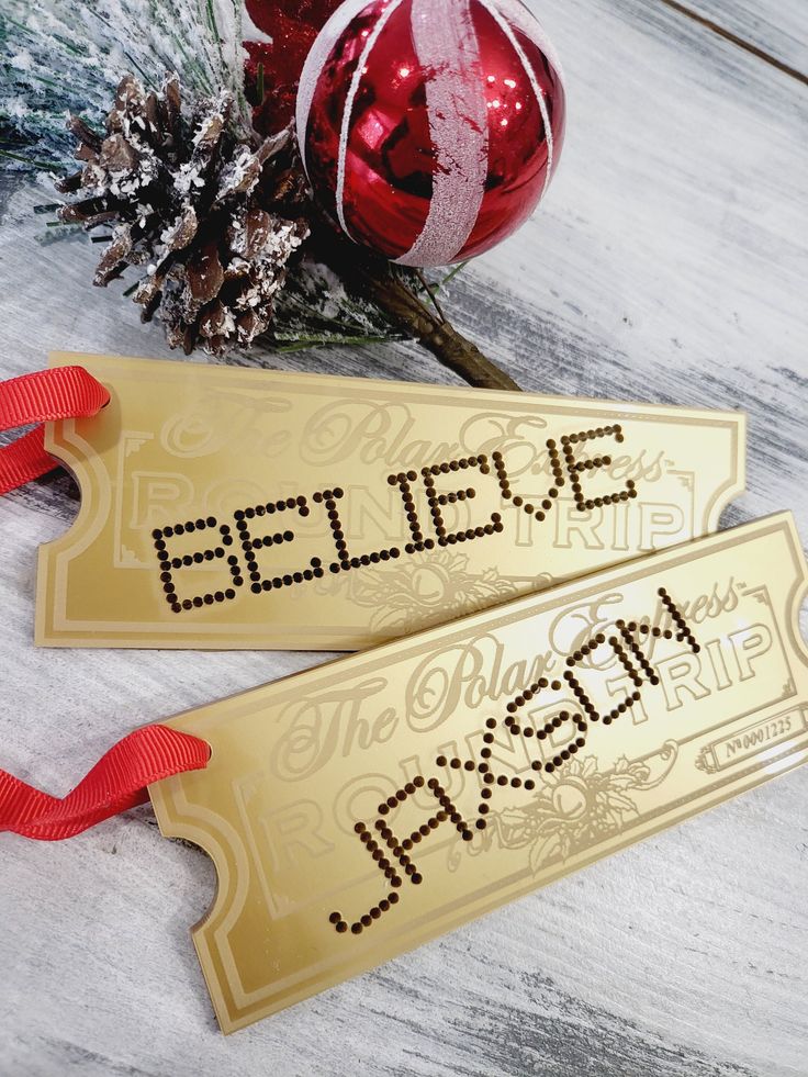 two gold christmas tags with the words believe and merry written on them next to a red ornament