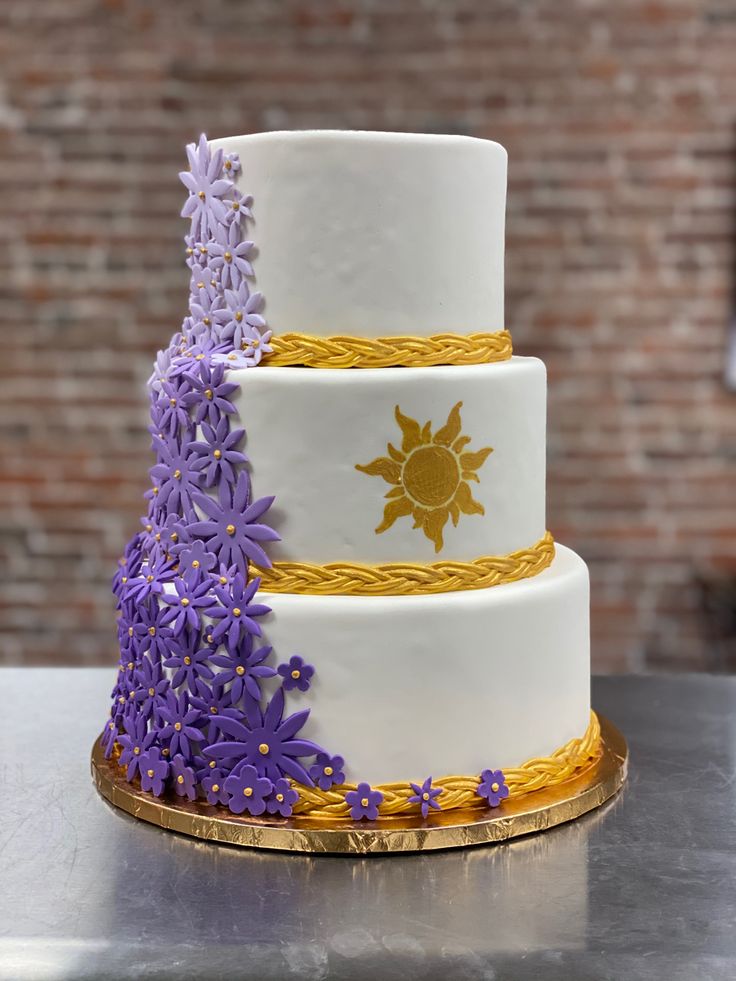 a three tiered cake with purple flowers on the bottom and gold trimmings