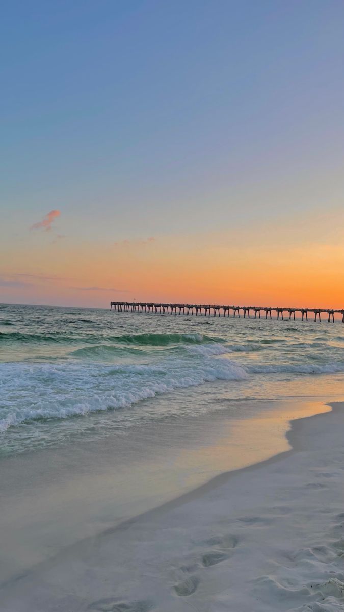 the sun is setting over the ocean with a long pier in the distance and waves crashing on the beach