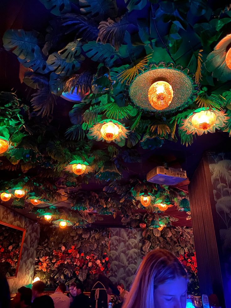 a woman sitting at a table in front of a bar with lights on the ceiling