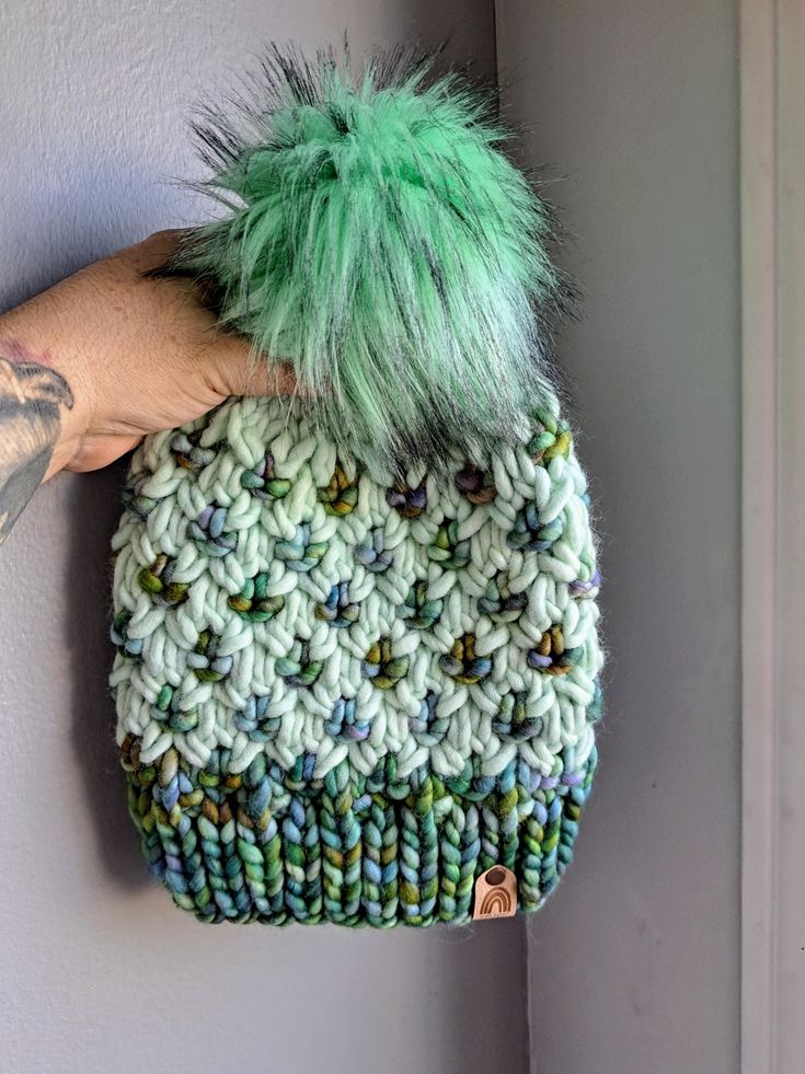 a hand holding a green and white knitted beanie with pom - pom