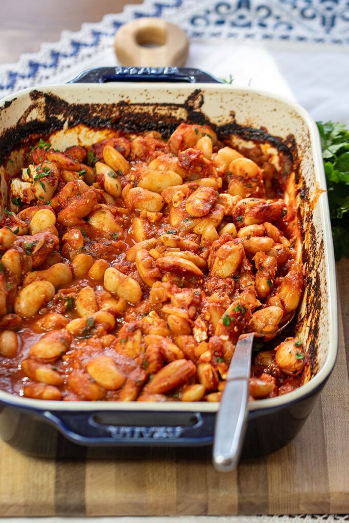 a casserole dish with beans and meat in tomato sauce on a wooden cutting board