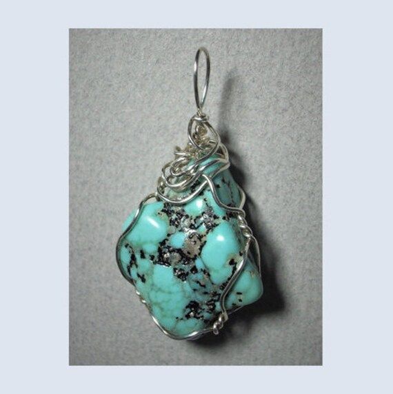 "Turquoise Nugget Pendant Wire Wrapped in .925 Sterling Silver Round Wire This inspiring stone offers a few small pebbles of golden pyrite nestled inside a light to medium turquoise-blue and black patterned background. Best worn with blue, green, coral, orange, or red; but quite versatile, and many dark, bright, pastel, and earth tone colors also wear well, especially black, grays, brown, beige/tan, and lavender. A similar color of turquoise will blend, so should be avoided. Some darker purple colors tend to clash. Take caution with yellows as well. Description: A light to bright turquoise-blue with black oxidation and very small pebbles of golden pyrite; rounded kite shaped, and opaque stone, wrapped in 22 gauge sterling silver wire. Stone Size: Medium (1-1/16\" x 7/8\" x 7/16\"). Pattern Artisan Turquoise Stones Necklace, Turquoise Chrysocolla Gemstone Necklace, Unique Wire-wrapped Turquoise Pendant Necklace, Unique Wire Wrapped Turquoise Pendant Necklace, Artisan Turquoise Necklace With Large Stone, Artisan Turquoise Necklace With Stones As Gift, Artisan Turquoise Stones Necklace For Gift, Unique Turquoise Gemstone Necklace, Distinctive Turquoise Gemstone Necklace