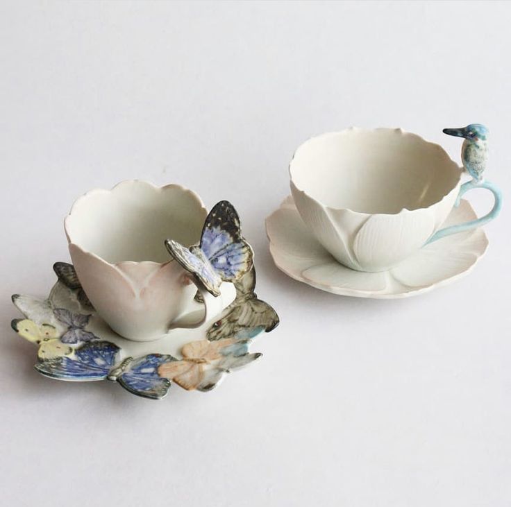 two porcelain cups and saucers decorated with flowers on white surface, one has a bird figurine in the middle