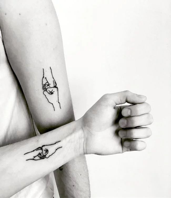 two hands holding each other with tattoos on their arms and wrist, one is pointing at the viewer