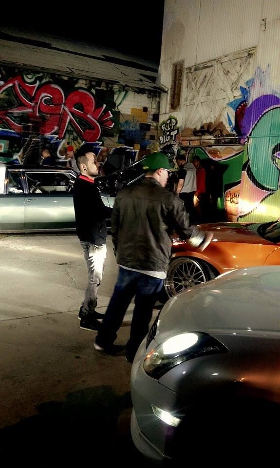 two men standing next to each other in front of parked cars and graffiti on the wall