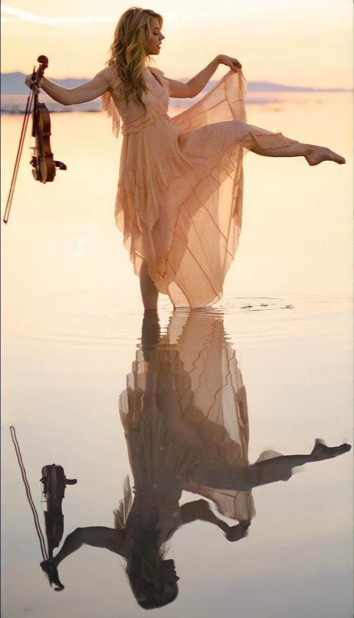 a woman in a dress is holding a violin and standing on the water's edge