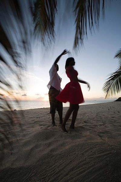 two people are dancing on the beach with palm trees in the foreground and water in the background