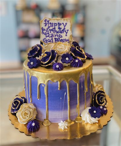 a blue and gold birthday cake with purple flowers on the bottom, sitting on a table