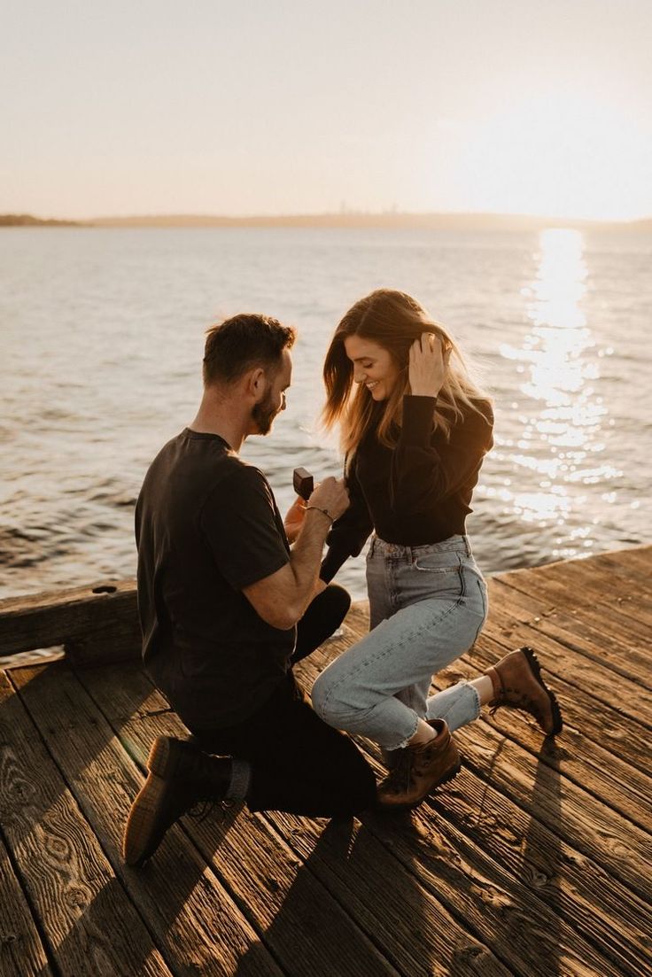 a man kneeling down next to a woman holding a glass of wine on top of a wooden pier