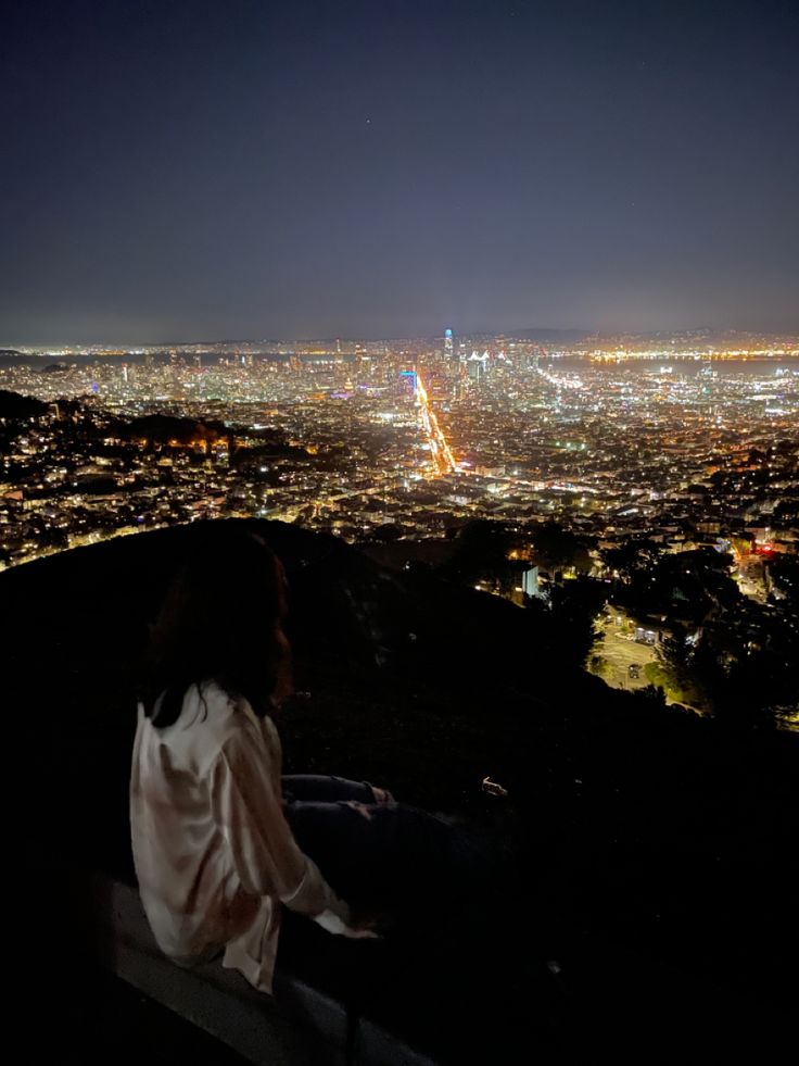 a woman sitting on top of a hill looking at the city lights