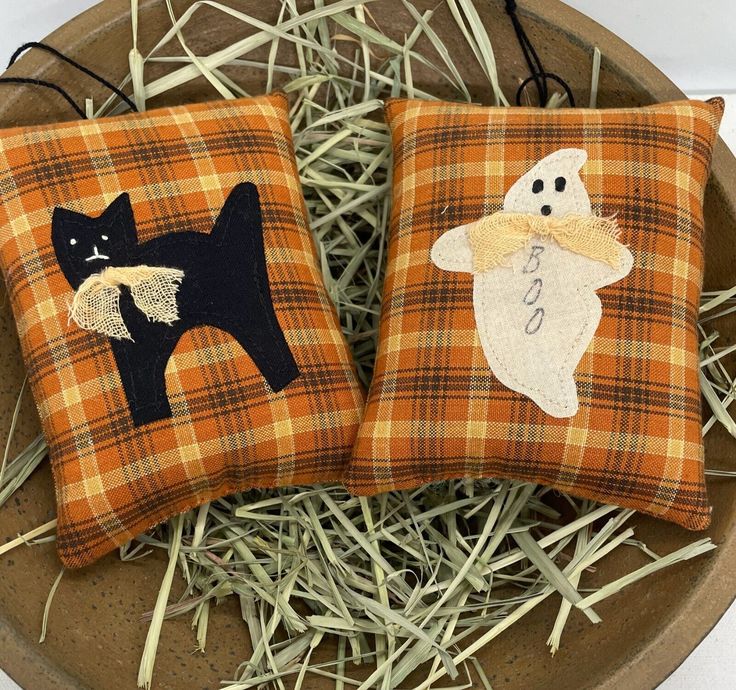 two decorative pillows with black cats and ghost on them sitting in some dry grass next to each other