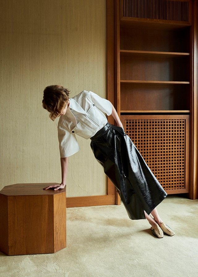 a woman leaning on a wooden block in front of a bookshelf and holding onto a black bag