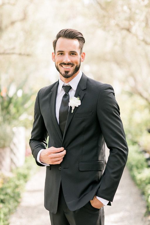 a man in a black suit and white flower boutonniere smiles at the camera