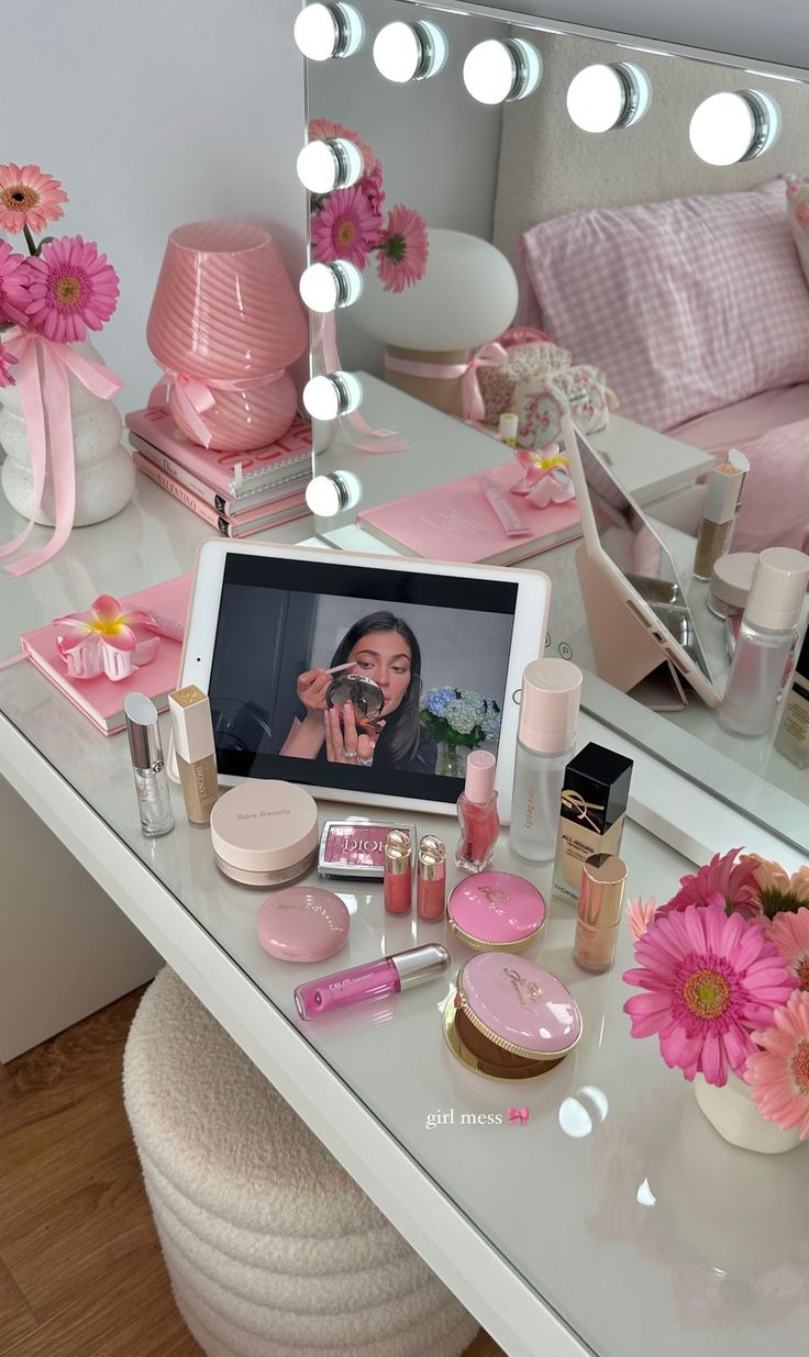 a vanity with pink flowers and makeup on it next to a mirror that is reflecting a woman's face