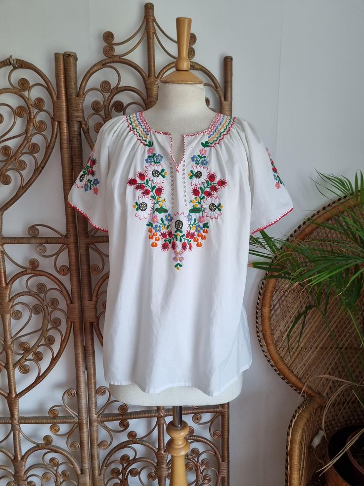 a white blouse with colorful flowers and beads on the collar is displayed in front of a chair