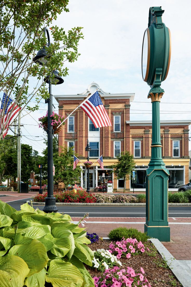 a green clock tower sitting on the side of a road next to flowers and trees