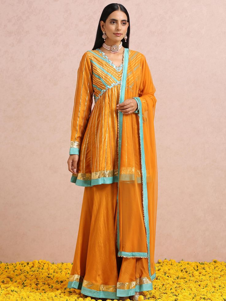 A three-piece Rust Orange Sharara set from the Label Nitika collection – Sunehra-Winter Festive 2022. Featured here is a beautiful rust orange peplum kurta in lightweight silk base paired with rust orange Sharara pants. The hand-embroidered floral teal sequins in symmetry along with teal-gota stripes on the V neck kurta gives an elegant touch to this gorgeous sharara set. This outfit is completed with a rust orange dupatta in net fabric bordered with gota embroidery edging. Orange Sharara, Embroidery Edging, Orange Dupatta, Pink Sharara, Gota Embroidery, Embroidered Sharara, Sharara Pants, Kurta Sharara Set, Silk Anarkali