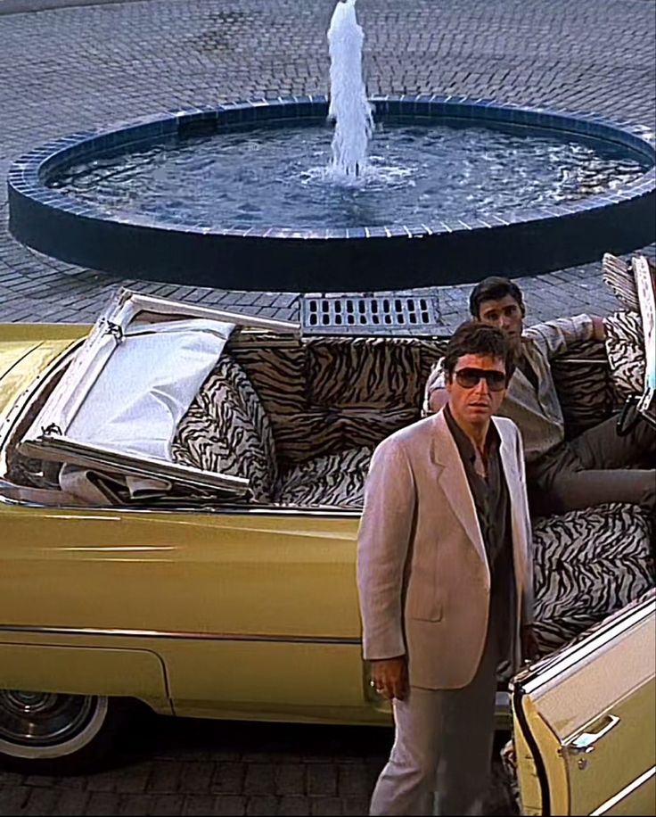 two men standing in front of a yellow car with luggage on the back and a fountain behind them