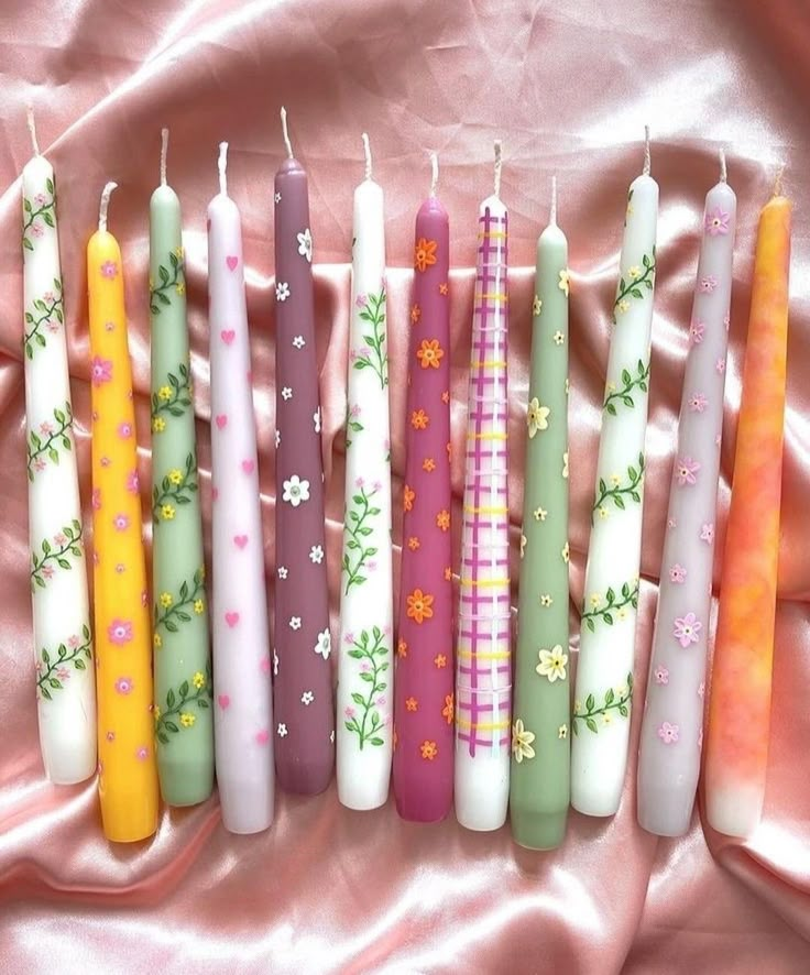 seven candles are lined up on a pink satin surface with daisies and flowers painted on them