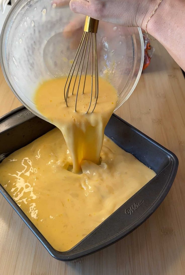 someone is mixing batter in a pan with a whisk on the side and an orange liquid pouring into it