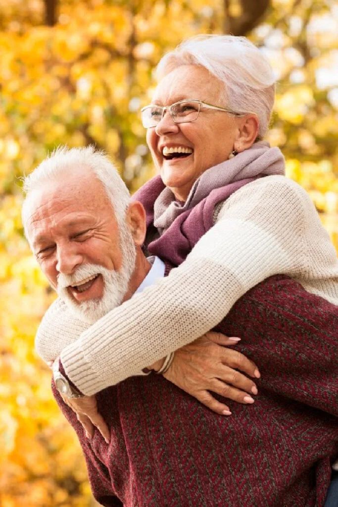 an older couple hugging each other in front of trees with autumn leaves on the ground