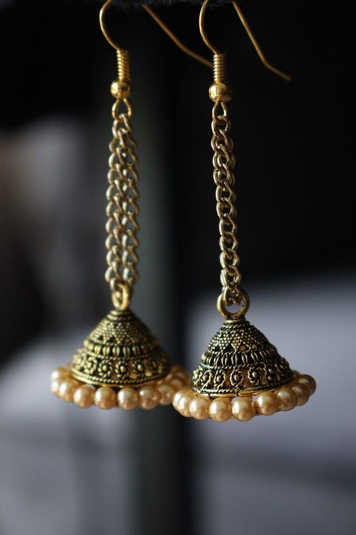 These earrings are a stunning pair of traditional jhumkas, characterized by their intricate design and antique finish. The earrings feature a delicate gold-toned chain, connecting to a jhumka adorned with vintage and ornate patterns. Surrounding the base of the jhumka is a row of small, round pearls, adding a touch of elegance and sophistication. The combination of the antique gold finish and the pearls creates a classic, timeless look, perfect for traditional occasions or to add a touch of eleg Brass Temple Jewelry Bridal Earrings With Latkans, Gold Brass Chandbalis For Diwali, Bollywood Style Gold Chandbalis In Brass, Brass Temple Jewelry Jhumkas For Festivals, Gift Brass Jhumkas With Latkans, Temple Jewelry Brass Jhumkas For Festivals, Gold Chandbali Jhumkas With Latkans, Diwali Brass Bridal Earrings With Latkans, Brass Bridal Earrings With Latkans For Diwali