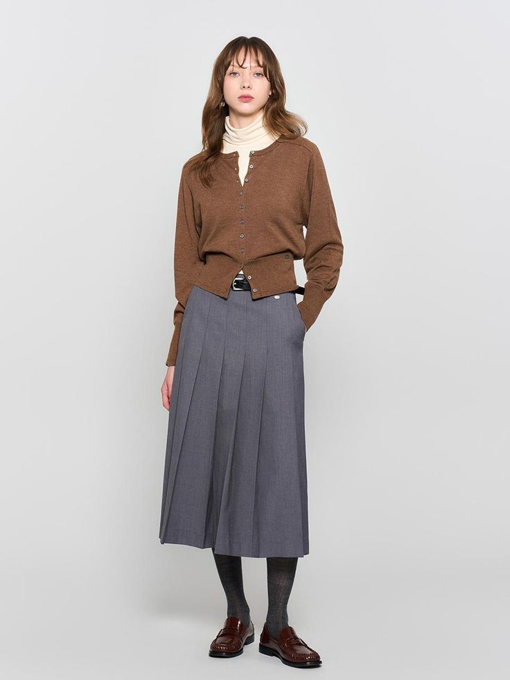 a woman standing in front of a white wall wearing a brown sweater and grey pleated skirt