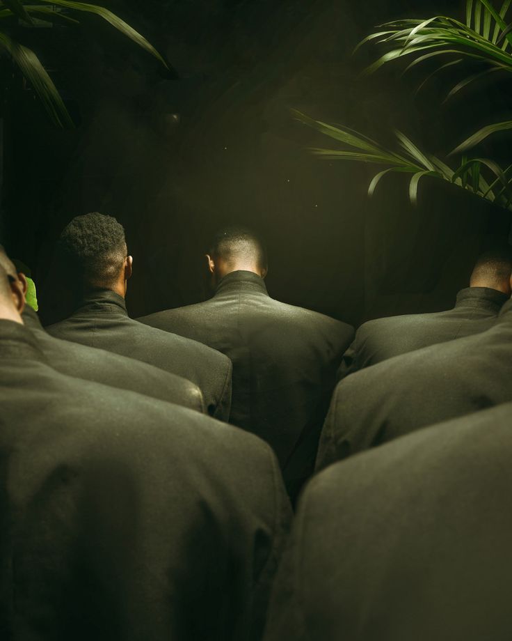 four men in black suits are walking through the dark tunnel with green plants on either side