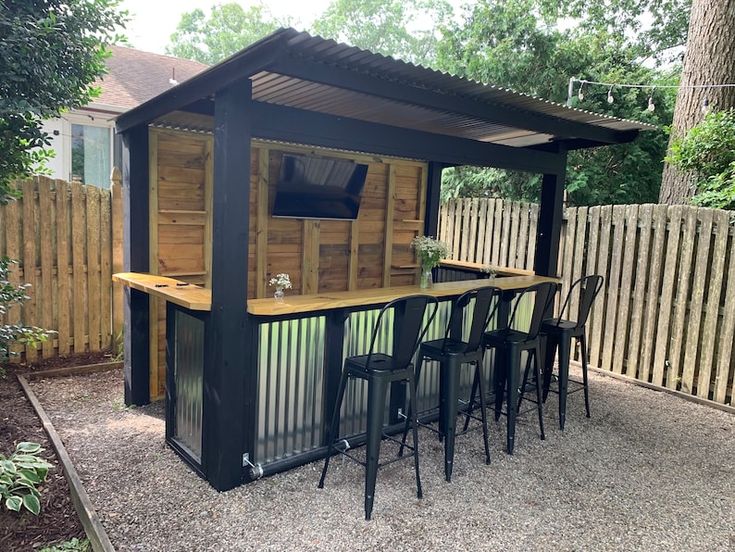 an outdoor bar with stools and tables in the middle of a backyard area next to a wooden fence