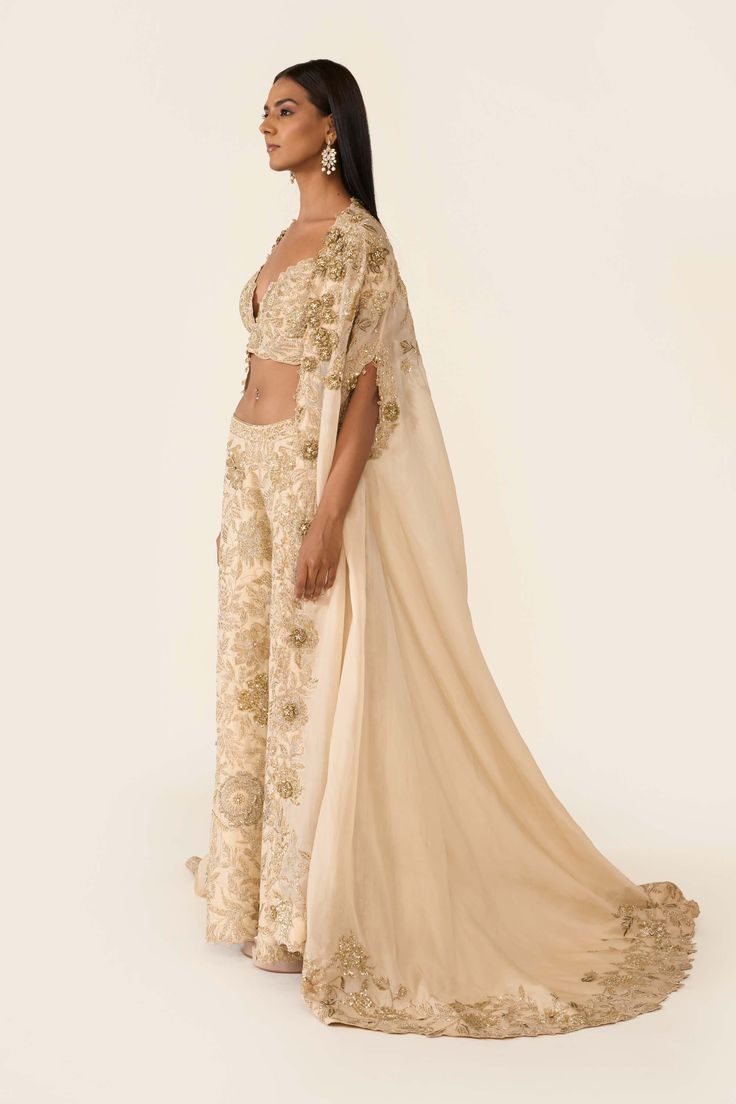 This golden cape is created using dori work, dabka, cutdana and sequins embellished with swarovski and 3d flowers, small crystals detailing on the edges. It is paired with golden blouse and trousers with thread and bead work. Blouse And Trousers, Golden Lehenga, Asian Couture, Varun Bahl, Dabka Work, Golden Blouse, Cape Blouse, Dori Work, Embroidered Cape